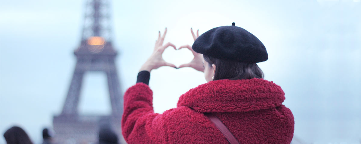 Femme de dos manteau rouge et béret basque noir faisant un cœur avec les doigts en face de la Tour Eiffel